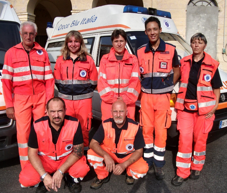 Gruppo di sette volontari di Croce Blu Italia in divisa arancione posano davanti a un'ambulanza bianca presso la sede di Asigliano Vercellese nel 2010