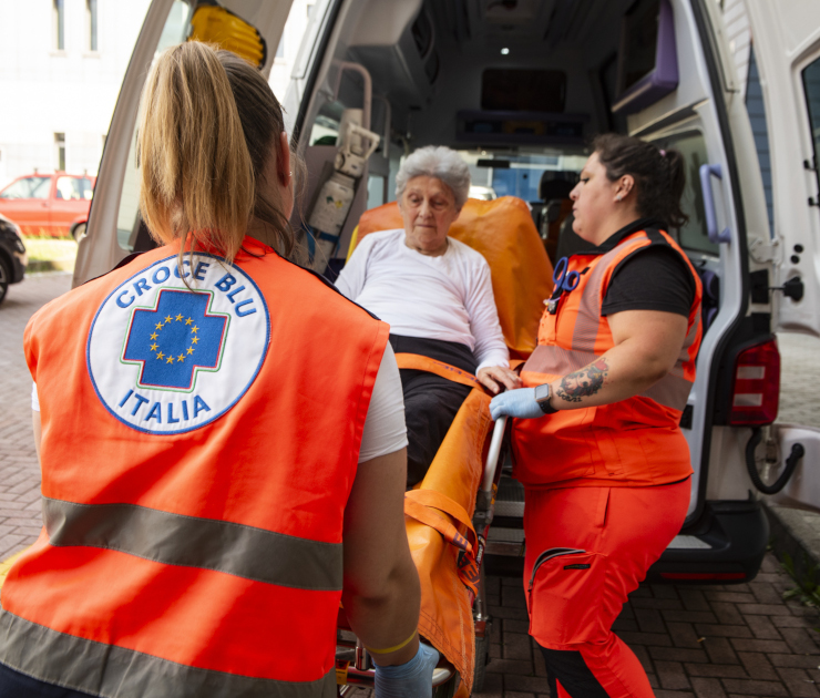 Due volontarie di Croce Blu Italia, che indossano pettorine arancioni, assistono un'anziana paziente seduta su una barella all'interno del vano di un'ambulanza.