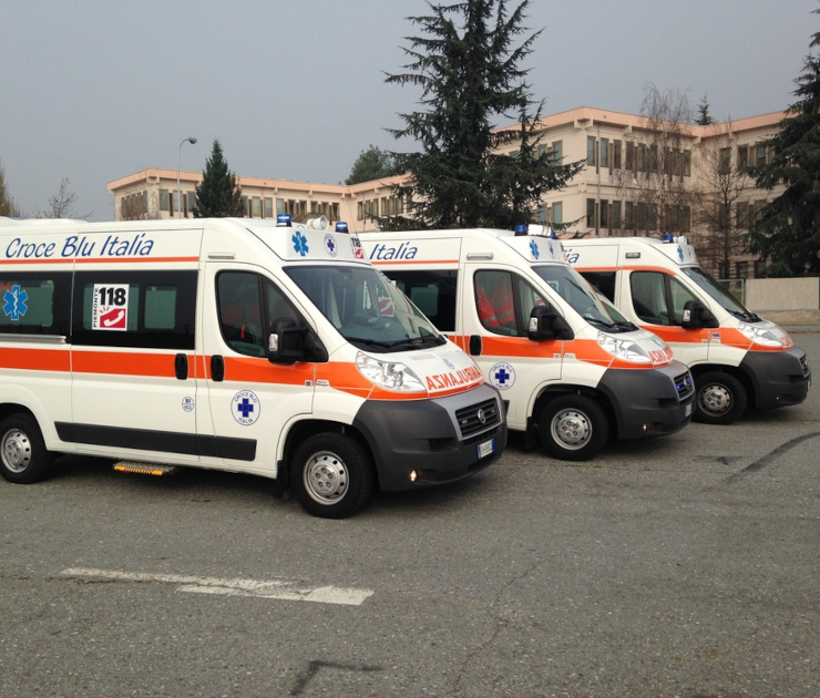La Flotta di Ambulanze di Croce Blu Italia Tre ambulanze di Croce Blu Italia (Fiat Ducato) allineate in sosta. I mezzi bianchi con la banda arancione e le scritte "118" e "Croce Blu Italia" sono parcheggiati davanti a un moderno edificio.