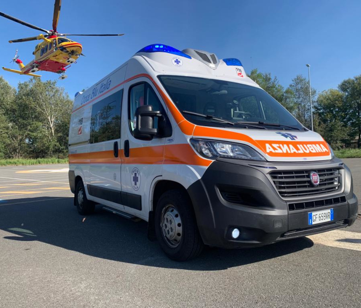 Ambulanza Mezzo di Soccorso Avanzato Croce Blu Italia Fiat Ducato bianca con bande arancioni e scritte 118, parcheggiata su eliporto con elicottero di soccorso giallo sullo sfondo. Mezzo attrezzato per emergenze con lampeggianti blu sul tetto.