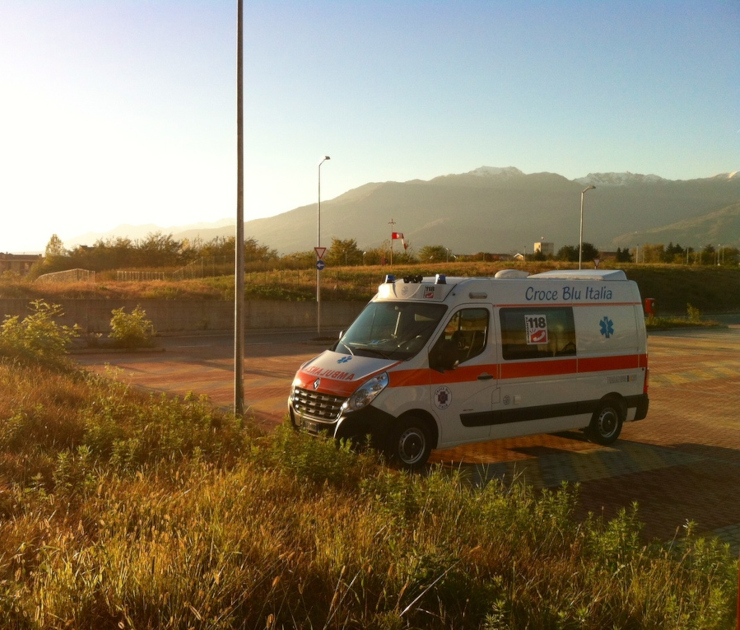 Ambulanza Renault Master bianca con banda arancione della Croce Blu Italia, con scritte 118 e stella della vita, fotografata all'alba in un parcheggio con montagne sullo sfondo.