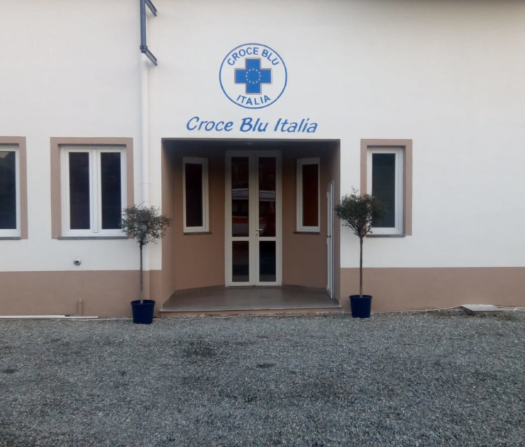 Facciata ingresso principale della sede Croce Blu Italia in Via Sermenza 24 a Vercelli dopo la ristrutturazione. Edificio bianco con zoccolatura beige, porta vetrata centrale, quattro finestre con cornici marroni, logo e scritta Croce Blu Italia sopra l'ingresso, due piante ornamentali in vasi blu ai lati dell'entrata, piazzale in ghiaia.