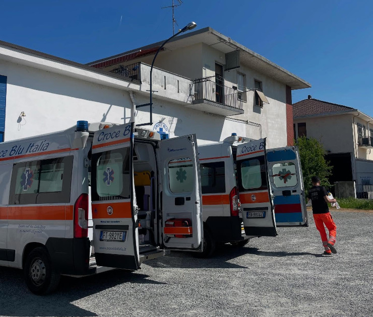 Sede operativa della Croce Blu Italia in Via Sermenza 24 a Vercelli: edificio bianco a due piani con scritta "Croce Blu Italia" sulla facciata, tre ambulanze Fiat Ducato bianche con banda arancione parcheggiate nel piazzale antistante, operatore in tuta rossa a destra.