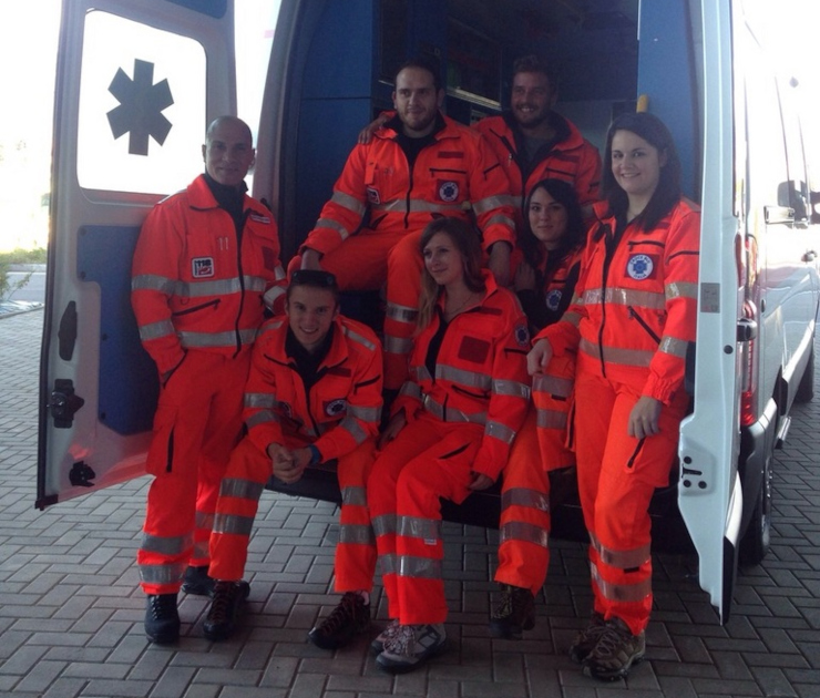 Gruppo di sette giovani volontari in divise ad alta visibilità rosse con bande rifrangenti seduti e in piedi davanti a un'ambulanza Croce Blu Italia. Foto di gruppo commemorativa per l'avvio del Servizio Civile Universale, gennaio 2020.
